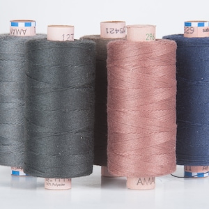 Op de afbeelding: Vier spoelen garen in grijs, donkergrijs, roze en blauw. De spoelen zijn gelabeld met de merknaam "AMAN" en het garennummer.