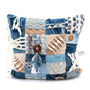 Op de afbeelding: Een patchwork denim tas met een verscheidenheid aan texturen en kleuren. De tas heeft vierkanten van blauwe denim, beige suède en crèmekleurig kant. Houten knopen en een veermotief voegen details toe. De tas heeft een macramé band.