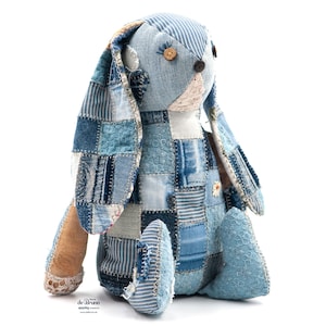 Könnte beinhalten: Eine Patchwork-Hasenpuppe aus blauem Denim. Der Hase hat Knopfaugen und einen Gänseblümchen-Akzent. Er besteht aus verschiedenen Denim-Flicken. Die langen, schlaffen Ohren und das Gesamtbild wirken skurril.