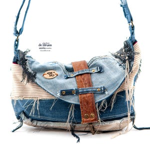 May include: A denim crossbody bag with a brown leather strap and buckle closure. The bag features a patchwork design with frayed edges and a striped fabric detail. The bag has a light blue denim flap and a wooden emblem.