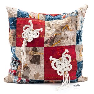 Puede incluir: Cojín cuadrado de patchwork con telas rojas, azules y beige. Cuenta con dos adornos de macramé blanco con cuentas de madera y borlas. El diseño combina texturas y patrones variados.