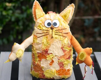 Può includere: Un peluche a forma di gatto realizzato con vari ritagli di tessuto arancione e giallo. La figura presenta grandi occhi circolari bianchi con pupille nere, baffi marroni ricamati e due braccia tese con cuscinetti gialli sulle zampe.