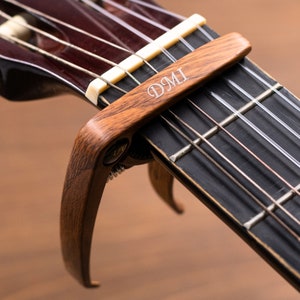 Könnte beinhalten: Nahaufnahme eines Holz-Kapodasters, der auf dem Griffbrett einer Gitarre befestigt ist. Der Kapodaster ist braun und trägt die Gravur "DMI". Die Saiten sind silberfarben, das Griffbrett dunkelbraun.