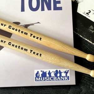 Peut inclure: Deux baguettes en bois avec le texte "Your Custom Text" imprim&eacute; dessus. Les baguettes sont pos&eacute;es sur une surface blanche avec un logo bleu qui dit "MUSICBANK".
