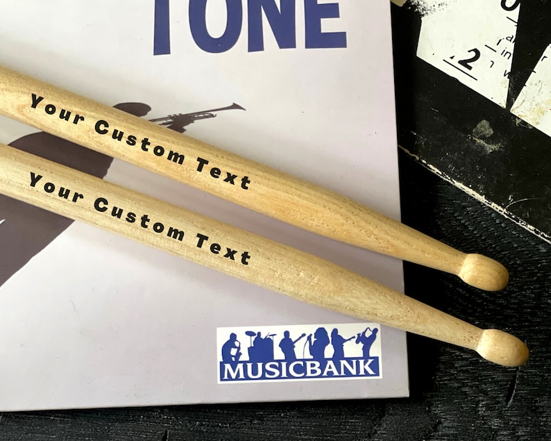 Peut inclure: Deux baguettes en bois avec le texte "Your Custom Text" imprim&eacute; dessus. Les baguettes sont pos&eacute;es sur une surface blanche avec un logo bleu qui dit "MUSICBANK".