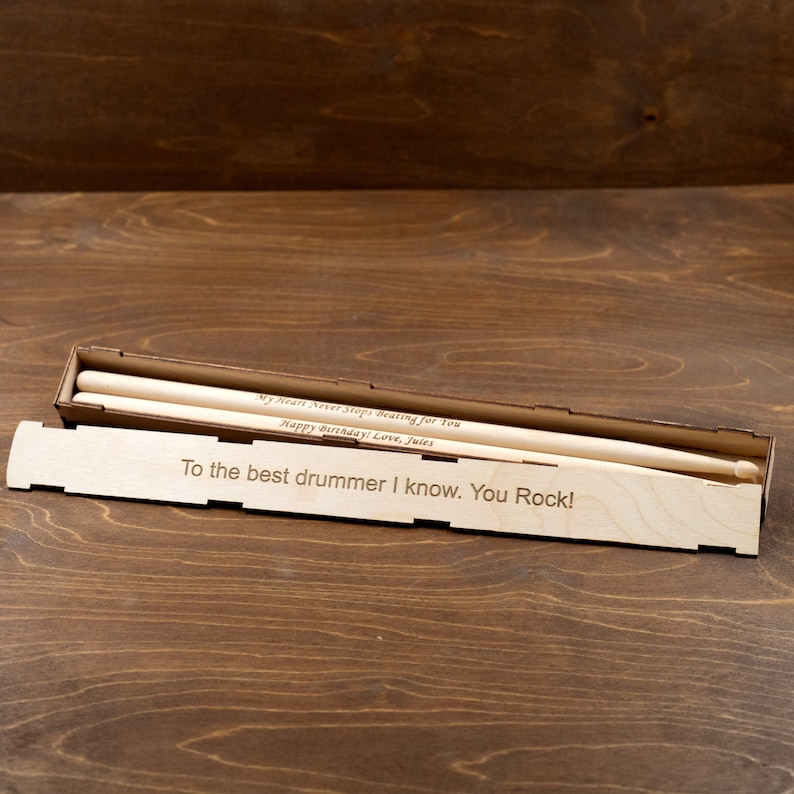 Peut inclure: Une bo&icirc;te en bois contenant deux baguettes de batterie en bois. La bo&icirc;te est grav&eacute;e avec le texte "My Heart Never Stops Beating for you Happy Birthday! Love, Jules" et "To the best drummer I know. You Rock!"