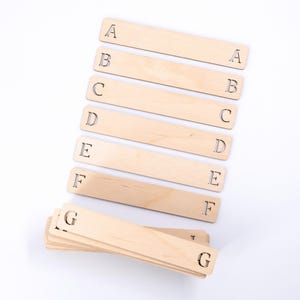 May include: Seven wooden bookmarks with letters A through G cut out. The bookmarks are stacked on top of each other, with the letter G on the top bookmark.