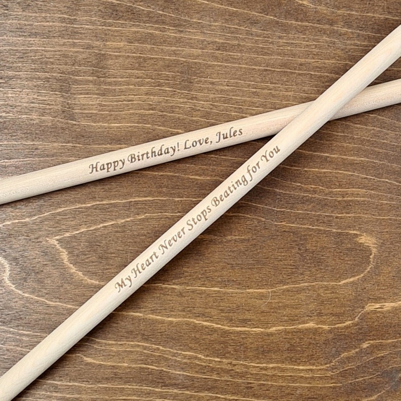 Peut inclure: Deux baguettes de batterie en bois avec le texte "Happy Birthday! Love, Jules" sur une baguette et "My Heart Never Stops Beating for You" sur l'autre baguette.