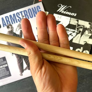 Peut inclure: Deux baguettes en bois tenues dans une main, avec une pochette d'album de disque en arri&egrave;re-plan. La pochette d'album pr&eacute;sente une image en noir et blanc de Louis Armstrong jouant de la trompette, avec le texte "Louis Armstrong" et "Weimar Jazz" visible.