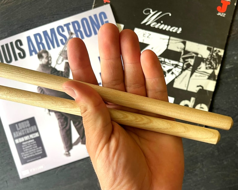 Peut inclure: Deux baguettes en bois tenues dans une main, avec une pochette d'album de disque en arri&egrave;re-plan. La pochette d'album pr&eacute;sente une image en noir et blanc de Louis Armstrong jouant de la trompette, avec le texte "Louis Armstrong" et "Weimar Jazz" visible.