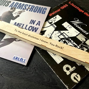 Peut inclure: Une bo&icirc;te en bois avec l'inscription "To the best drummer I know. You Rock!" La bo&icirc;te est pos&eacute;e sur deux disques vinyles. Un disque est un album de Louis Armstrong intitul&eacute; "In A Mellow" et l'autre est un album Jazztage der DDR intitul&eacute; "Weimar 1985".