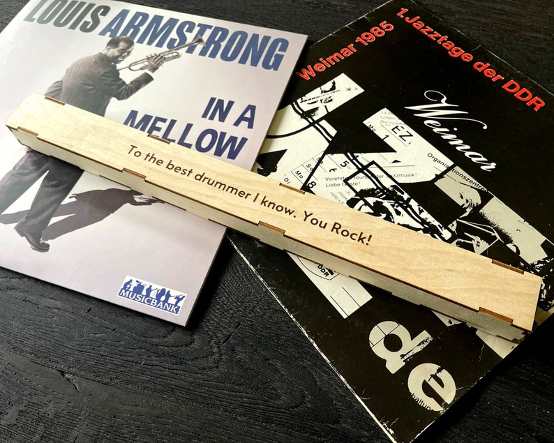 Peut inclure: Une bo&icirc;te en bois avec l'inscription "To the best drummer I know. You Rock!" La bo&icirc;te est pos&eacute;e sur deux disques vinyles. Un disque est un album de Louis Armstrong intitul&eacute; "In A Mellow" et l'autre est un album Jazztage der DDR intitul&eacute; "Weimar 1985".