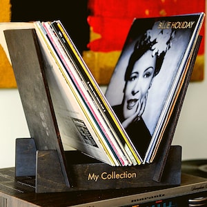 Puede incluir: Un soporte para discos de madera oscura con un disco de vinilo negro apoyado en él. La portada del disco presenta una foto en blanco y negro de Billie Holiday. El soporte tiene las palabras "My Collection" grabadas en la parte delantera.