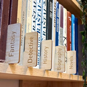 May include: Wooden bookends labeled "Fantasy", "Detective", "History", "Biography", and "Novels" stand between books on a shelf. The bookends are light brown with dark brown text. The books have various colors and titles.