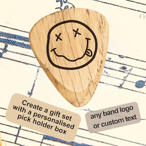 Peut inclure: Un médiator de guitare en bois avec un smiley noir aux yeux en 'X' et la langue tirée. Le médiator est posé sur une partition de musique avec le texte 'Create a gift set with a personalised pick holder box' et 'any band logo or custom text'.