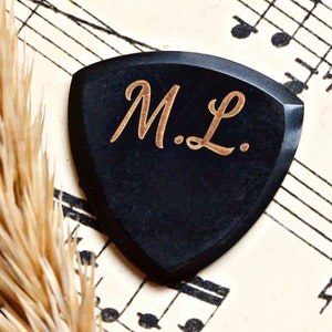 May include: A black guitar pick with a gold inscription that reads "M.L.". The pick is lying on a sheet of music.