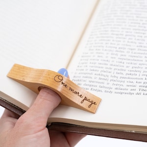 Puede incluir: Un sujetapáginas de madera clara con el texto "One more page" grabado. El soporte está diseñado para sujetarse con un dedo. El soporte está colocado en un libro abierto.