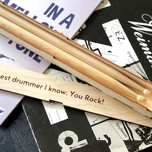 Peut inclure: Bo&icirc;te en bois contenant deux baguettes de batterie en bois. La bo&icirc;te porte la gravure "the best drummer I know. You Rock!" sur le devant.