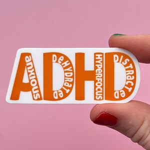 Op de afbeelding: Een witte sticker met het woord "ADHD" in grote oranje letters. Kleinere oranje woorden zoals "angstig", "uitgedroogd", "hyperfocus" en "afgeleid" staan ook op de sticker. De sticker wordt tegen een roze achtergrond gehouden.