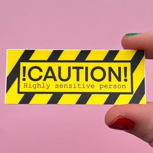 Może przedstawiać: Żółta prostokątna naklejka z czarnymi ukośnymi paskami i napisem "!CAUTION!" pogrubioną czcionką. Poniżej znajduje się napis "Highly sensitive person". Naklejka jest trzymana na różowym tle.