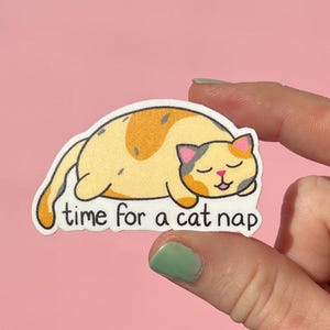 May include: A sticker featuring a cartoon cat in a sleeping pose. The cat is yellow with orange spots and gray accents. The text "time for a cat nap" is below the cat. The sticker is held against a pink background.