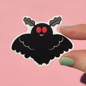 May include: A black moth sticker with a white outline, red eyes, and small antennae. The sticker has a cartoon-like design and is held against a pink background.