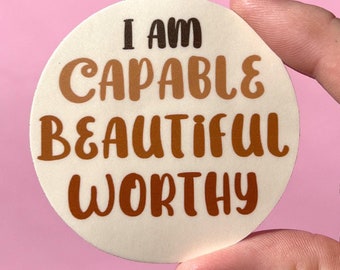 May include: A round sticker with the text "I am capable beautiful worthy" in brown and black lettering on a cream background.