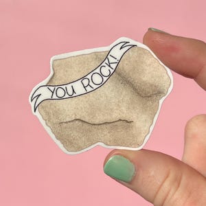 May include: A sticker featuring a beige rock shape with a white banner that reads "You Rock!" in black text. The sticker has a white border and is held by a person with a pink background.