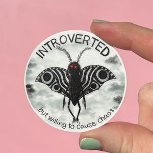 May include: A round sticker featuring a black moth with red eyes and patterned wings, set against a cloudy background. The text "INTROVERTED" arches above the moth, with "but willing to cause chaos" below.