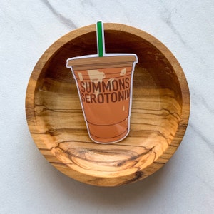 May include: A sticker of a brown iced coffee cup with a green straw and the text "SUMMONS SEROTONIN" on a wooden surface.