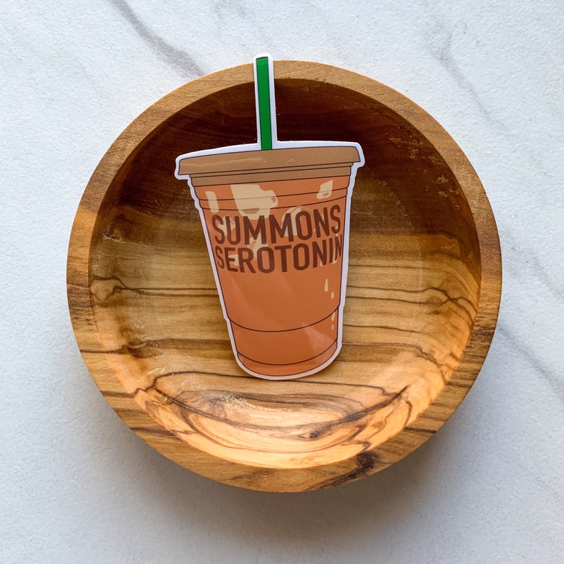 May include: A sticker of a brown iced coffee cup with a green straw and the text "SUMMONS SEROTONIN" on a wooden surface.