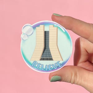 May include: A round sticker featuring a stylized illustration of a building with a central tower and the word "BELIEVE" in a blue, bubble-style font. The sticker is set within a bubble-like design with a light blue and teal gradient.