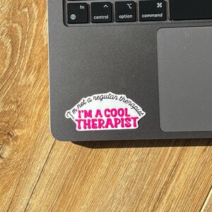May include: A white sticker with black text that reads "I'm not a regular therapist I'M A COOL THERAPIST" in a pink, curved font. The sticker is on a grey laptop.