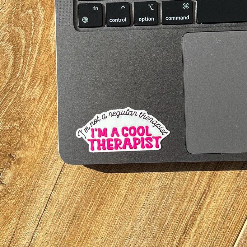 May include: A white sticker with black text that reads "I'm not a regular therapist I'M A COOL THERAPIST" in a pink, curved font. The sticker is on a grey laptop.