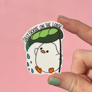 May include: A white sticker featuring a cartoon goose under a green leaf umbrella, with blue raindrops. The text "SILLY GOOSE ON THE LOOSE" is arched above the goose. The goose has orange feet and a beak.