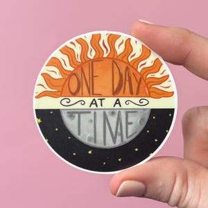 May include: A round sticker with a sun and moon design. The sun is orange and yellow with rays. The moon is gray with a black background and stars. The text "ONE DAY AT A TIME" is written in brown on the sticker.