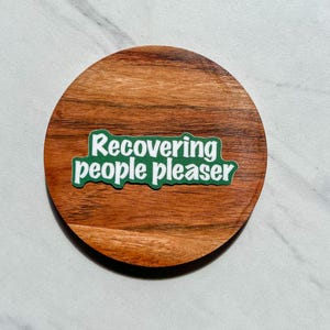 May include: A green sticker with white text that reads "recovering people pleaser" on a wooden coaster.