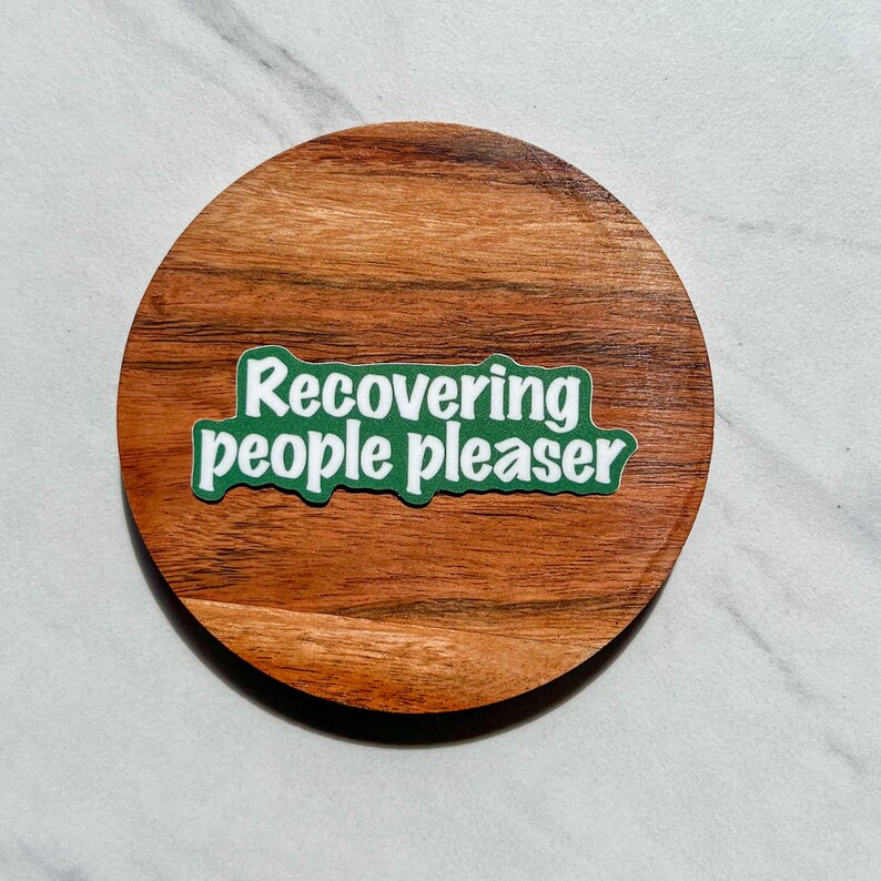 May include: A green sticker with white text that reads "recovering people pleaser" on a wooden coaster.