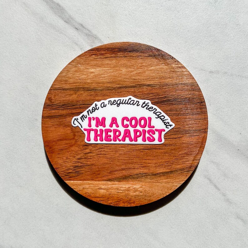 May include: A white sticker with pink text on a wooden coaster. The sticker says "I'm not a regular therapist I'm a cool therapist."