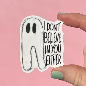 May include: A white ghost-shaped sticker with black eyes and a black outline. The sticker has the text "I DON'T BELIEVE IN YOU EITHER" in black. The sticker is held against a pink background.
