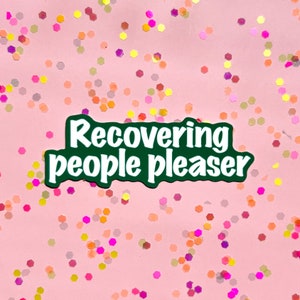 May include: Green sticker with white text that reads "recovering people pleaser" on a pink background with colorful glitter.