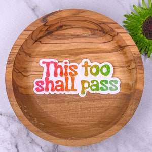 May include: A white sticker with the text "This too shall pass" in rainbow colours with white polka dots on a wooden surface.