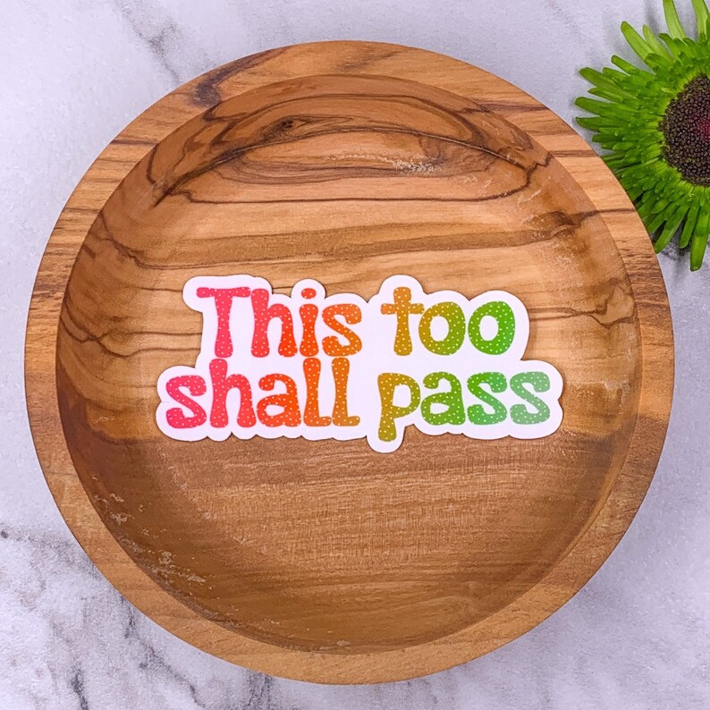 May include: A white sticker with the text "This too shall pass" in rainbow colours with white polka dots on a wooden surface.