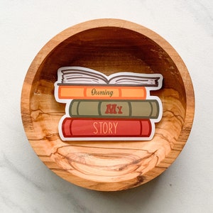 May include: A sticker of a stack of books with the text "Owning My Story" on a wooden bowl.