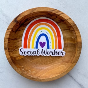 May include: A rainbow sticker with a purple heart in the centre and the text "Social Worker" below. The sticker is on a wooden surface.