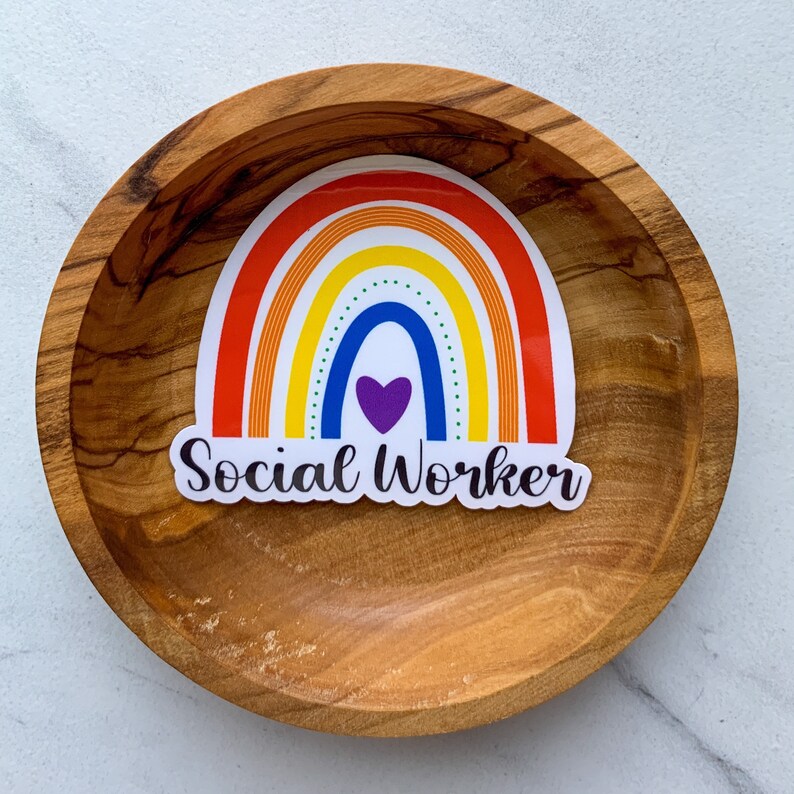 May include: A rainbow sticker with a purple heart in the centre and the text "Social Worker" below. The sticker is on a wooden surface.