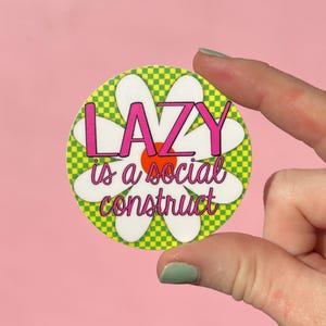 May include: A round sticker with a daisy design. The center is orange, surrounded by white petals. The background is a green and yellow checkerboard pattern. The text "LAZY is a social construct" is in pink and white.