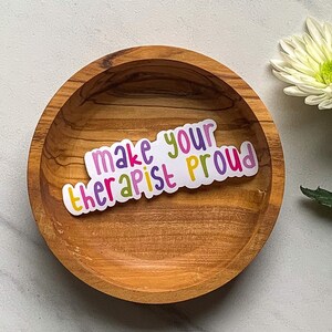 May include: A sticker with the text "make your therapist proud" in a rainbow of colours. The sticker is on a wooden bowl.