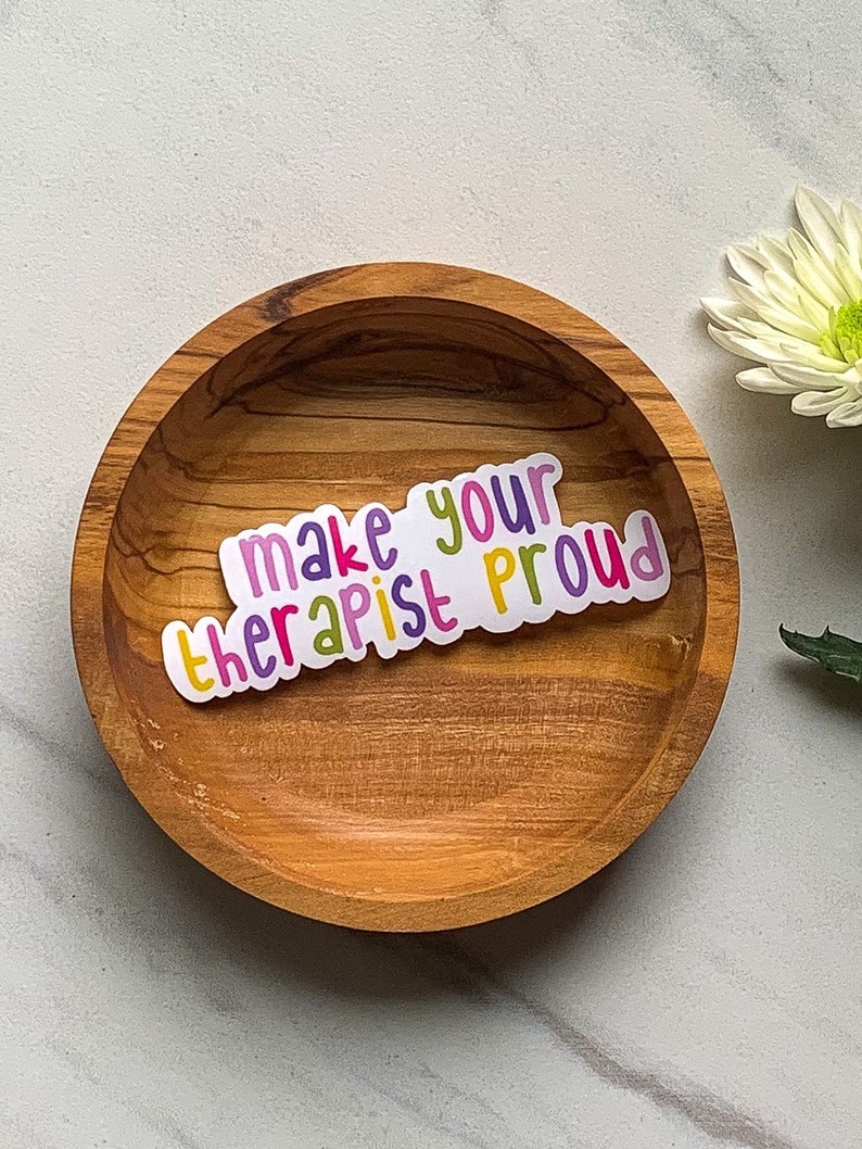 May include: A sticker with the text "make your therapist proud" in a rainbow of colours. The sticker is on a wooden bowl.