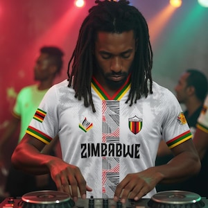 May include: A white jersey with the word "ZIMBABWE" in black, featuring a geometric design in black and red. The jersey has green, yellow, and red accents, and a Zimbabwe flag patch on the sleeve. The person is wearing dreadlocks.